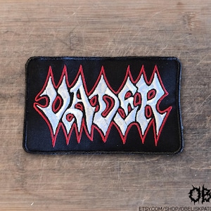 May include: A rectangular black embroidered patch featuring the word VADER in a stylized, jagged white font with a thin red outline. The embroidery is dense, creating a textured surface on the fabric, with sharp, pointed edges on each letter.