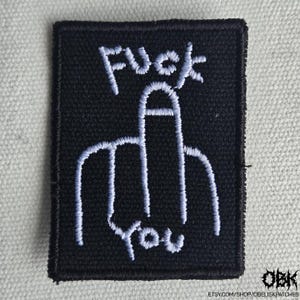 Puede incluir: Parche bordado negro con las palabras "FUCK YOU" y un gesto con la mano en hilo blanco. El parche es rectangular y tiene un borde cosido. El nombre de la tienda Etsy es visible en la esquina inferior derecha.
