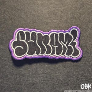 May include: Embroidered patch with the word "SBASK" in a bubble letter style. The letters are black with a white outline, set against a black background, and framed by a purple border. The patch is on a dark gray surface.