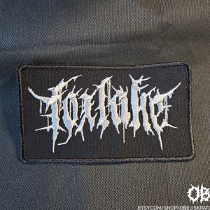 May include: A rectangular black embroidered patch featuring the white text Foxlake in a stylised, jagged, and sharp-edged font. The patch has a black stitched border and is set against a dark, textured fabric background with a small white logo in the corner.
