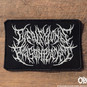 May include: A rectangular black embroidered patch featuring white text in a stylized, jagged, and thorny font that reads THE FISTION PRISONPLANET. The patch has a thick black stitched border and is designed for attachment to clothing or accessories.