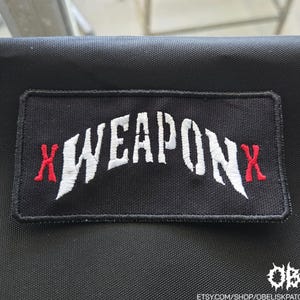 以下が含まれることがあります： 中央に白い文字でWEAPONとアーチ状に刺繍され、両端に赤い文字Xが配置された長方形の黒い刺繍ワッペン。ワッペンには黒いステッチの縁取りがあり、黒い布地の上に置かれています。