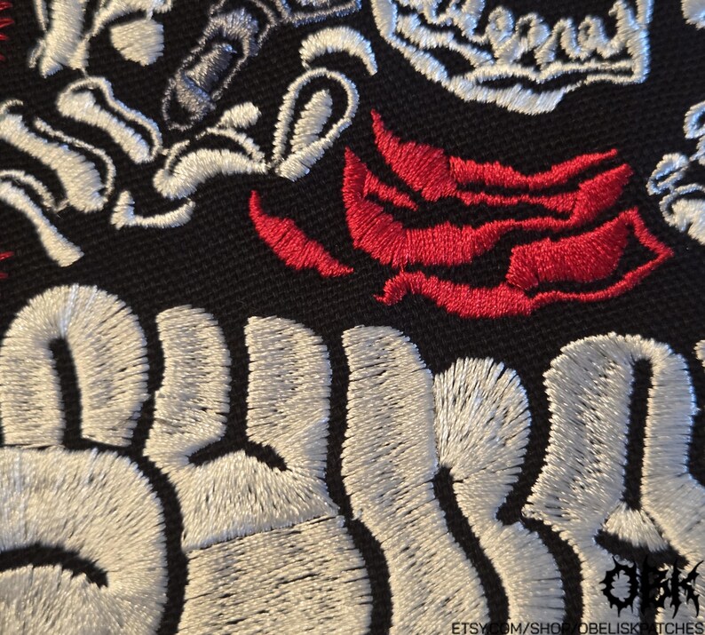K&ouml;nnte beinhalten: Ein schwarzer Stoffaufn&auml;her mit wei&szlig;er Stickerei in Form von Buchstaben und knochenartigen Elementen, akzentuiert durch rote, bl&uuml;tenblatt- oder flammenartige Stickereien. Der Text ist in einer fetten, stilisierten Schrift teilweise sichtbar.