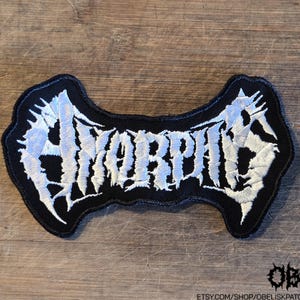 May include: A black embroidered patch featuring the word Amorphis in white, jagged, stylized lettering. The patch has a distinct, irregular shape that follows the contours of the text, with a thick black border surrounding the white embroidery.