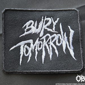 May include: A rectangular black embroidered patch featuring the white text 'BURY TOMORROW' in a stylised, jagged font. The patch has a thick black stitched border and a textured fabric base, with a small white logo in the bottom right corner.