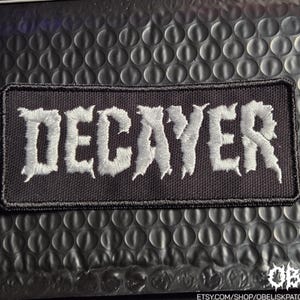 May include: A rectangular embroidered patch with a black fabric background and white stitched lettering that reads DECAYER. The text features a jagged, distressed font style with a thick black border surrounding the entire rectangular shape.