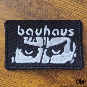 May include: A rectangular black embroidered patch featuring the word "bauhaus" in white lowercase letters above a stylised white graphic design. The patch has a thick black stitched border and a textured fabric base, with clear white thread embroidery.