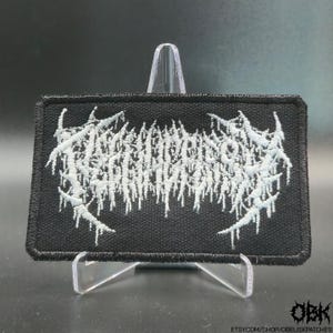 May include: Black rectangular patch with white embroidered text. The text has a dripping, stylized design. The patch is displayed on a clear stand. The bottom right corner of the patch has the letters "OBK" and the Etsy shop URL.