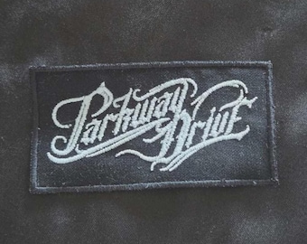 May include: A rectangular black fabric patch featuring the white embroidered text Parkway Drive in a stylized, cursive script font. The patch has a stitched border and is set against a solid black background, displaying clear, detailed embroidery.