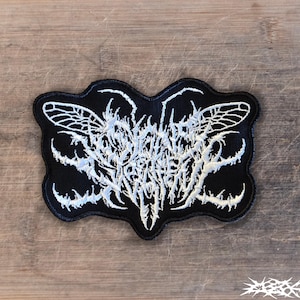 May include: A black embroidered patch featuring white thread in a complex, jagged, and illegible script style. The design incorporates insect-like wing shapes on the sides and sharp, thorn-like protrusions extending from the central text area.