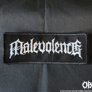 May include: A rectangular black embroidered patch featuring the word Malevolence in white gothic-style lettering. The patch has a stitched border and is set against a black fabric background. The text OBK is visible in the bottom right corner.