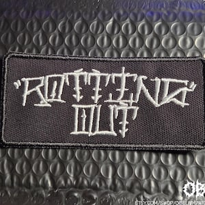 May include: A rectangular black embroidered patch featuring the white text ROTTING OUT in a stylized, sharp-edged font. The patch has a thick black stitched border and a textured fabric surface, designed for application onto various textile items.