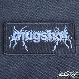 May include: A rectangular black fabric patch featuring the word "mugshot" embroidered in a light blue, jagged, stylized font. The lettering has sharp, branch-like extensions reaching outward from the characters, creating a distressed appearance.
