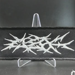 May include: A rectangular black fabric patch featuring an embroidered design in white thread. The embroidery depicts a series of sharp, interconnected, thorn-like or barbed wire shapes arranged in a horizontal, abstract cluster across the center.