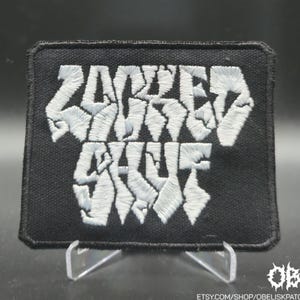 May include: Black rectangular patch with the words "ZOMBIE SKULL" embroidered in white. The patch is displayed on a clear stand against a dark background. The Etsy shop URL is visible at the bottom.