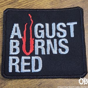 以下が含まれることがあります： 黒色の刺繍パッチ。白文字で「AUGUST BURNS RED」と赤色の炎のデザイン。パッチは黒い縁取りで長方形です。衣類やバッグの装飾に最適です。