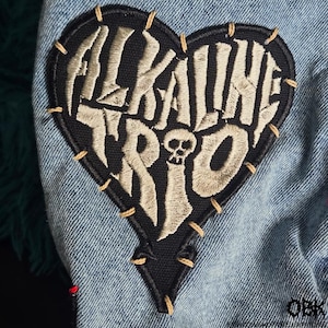 Könnte beinhalten: Ein herzförmiger Aufnäher mit den Worten "ALKALINE TRIO" in einer stilisierten Schriftart, mit einer kleinen Totenkopf-Grafik. Der Aufnäher ist schwarz mit beigefarbener Stickerei und auf einem Jeansstoff mit beigem Faden aufgenäht.