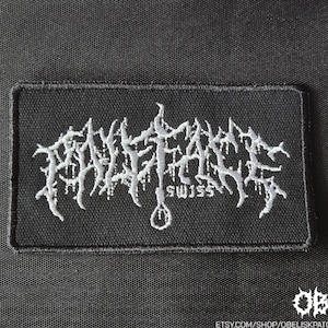 May include: Black rectangular patch with white embroidered text. The text is stylized and reads "PHALLIC FAECES SWISS". A small white graphic of a noose is below the text. The patch is on a black background.