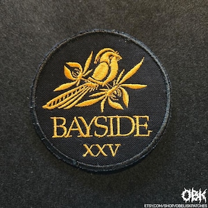 May include: A circular black embroidered patch featuring a gold-colored bird perched on a branch with leaves. The text BAYSIDE is embroidered in gold below the bird, with the Roman numerals XXV positioned underneath the text in gold thread.