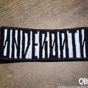May include: Black and white embroidered patch with the word "UNEVEN" in a bold, stylized font. The text is white against a background of vertical black stripes. The patch has a black border and is rectangular in shape.