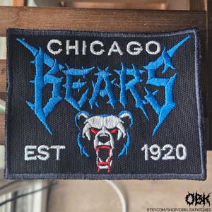 May include: Black rectangular patch with the words "CHICAGO BEARS" in white and blue, and "EST 1920". A stylized bear head with red eyes and mouth is in the center. The patch is likely for clothing or accessories.