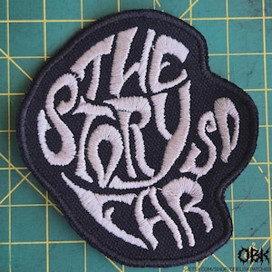 May include: Black embroidered patch with the words "THE STORY SO FAR" in a stylized, light gray font. The patch has a unique, irregular shape and is likely designed to be sewn onto clothing or accessories.