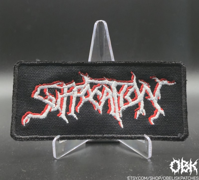 May include: Black rectangular patch featuring the word "SUFFOCATION" in red and white embroidery. The patch is displayed on a clear plastic stand. The background is dark. The patch is likely for a jacket or bag.