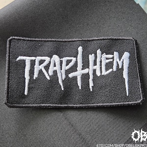 May include: A rectangular black embroidered patch featuring the white text TRAP THEM in a stylised, jagged font. The patch has a black stitched border and a textured fabric surface, with the text clearly centred across the middle of the design.