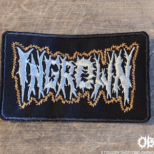 May include: A rectangular black embroidered patch featuring the word INGROWN in stylized, jagged, light gray lettering. The text is outlined with a thin, irregular border of orange thread, and the entire patch has a black stitched border.