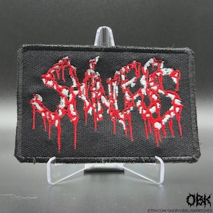 May include: Black rectangular patch with the word "SNAKES" embroidered in white and red. The red embroidery has a dripping effect. The patch is displayed on a clear stand.