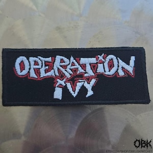 May include: A rectangular black patch embroidered with the words "OPERATION IVY" in white, outlined in red. A red starburst design is visible within the text. The patch is likely for clothing or accessories.