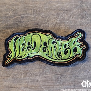 May include: An embroidered patch featuring the text "WEED EATER" in a stylised, flowing lime green font with a black background and a thin orange border. The lettering has a jagged, organic appearance with sharp, pointed edges throughout the design.