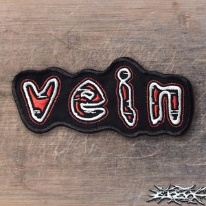 May include: A black embroidered patch featuring the word VEIN in stylized lettering. The letters are filled with red thread and outlined in thick white stitching, set against a black fabric background with a black stitched border edge.