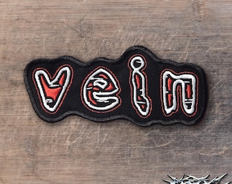 May include: A black embroidered patch featuring the word VEIN in stylized lettering. The letters are filled with red thread and outlined in thick white stitching, set against a black fabric background with a black stitched border edge.