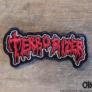 May include: An embroidered patch featuring the text TERRORIZER in a stylized, dripping red font with a white outline on a black background. The lettering has a jagged, distressed appearance, and the patch has a black border following the shape of the text.