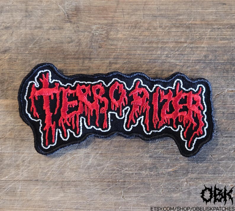 Puede incluir: Un parche bordado que presenta el texto TERRORIZER en una fuente roja estilizada y goteante con un contorno blanco sobre un fondo negro. Las letras tienen una apariencia irregular y desgastada, y el parche tiene un borde negro que sigue la forma.