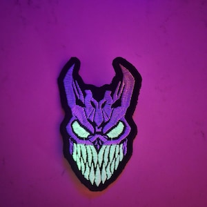 May include: Embroidered patch depicting a purple demonic face with glowing green eyes and teeth, set against a black border. The patch is designed for application to clothing or accessories. The design is detailed and stylized.