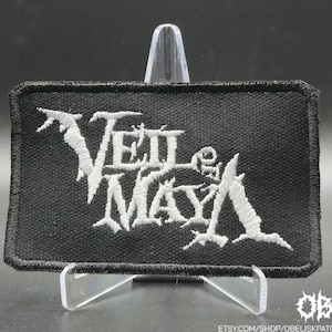 May include: Black rectangular patch with the band name "Veil of Maya" embroidered in white. The patch has a black border and is displayed on a clear stand. The band's logo is in a gothic style.