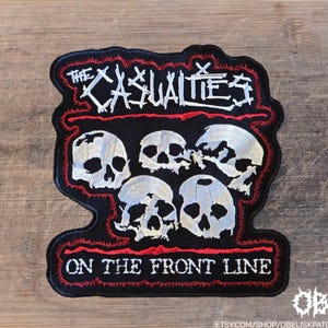 May include: A black embroidered patch featuring the white text THE CASUALTIES at the top and ON THE FRONT LINE at the bottom. The center displays five white skulls arranged in a cluster, all outlined with a jagged red stitched border.