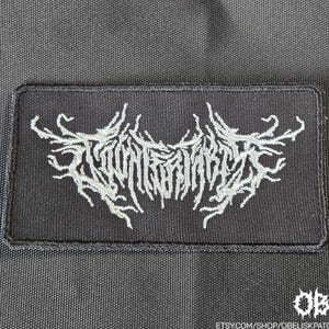 May include: A rectangular black fabric patch featuring white embroidered text in a stylized, jagged, branch-like font. The embroidery is centered on the patch, which has a stitched border. In the bottom right corner, the text OBK is visible.