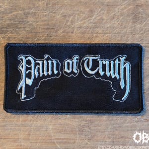 May include: A rectangular black fabric patch featuring the white embroidered text Pain of Truth in a stylized gothic font. The patch has a black stitched border and a textured surface, with the text centered across the middle of the design.