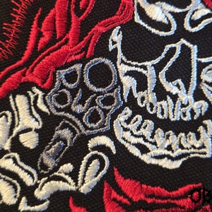 K&ouml;nnte beinhalten: Ein schwarzer Stoffaufn&auml;her mit detaillierter Stickerei eines wei&szlig;en Totenkopfes und einer Skeletthand, die einen Revolver h&auml;lt, akzentuiert durch leuchtend rote Flammenmuster. Unten rechts steht der Text "OBK" und "ETSY.COM/SHOP/OBELISKPATCHES".