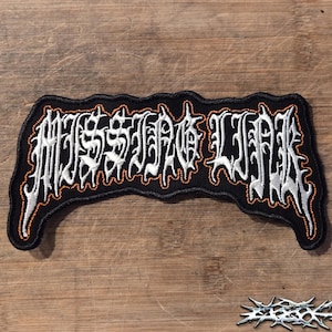 May include: A black embroidered patch featuring the text MISSING LINK in a stylized, jagged white font with an orange border. The patch has an irregular, wavy shape and is designed with a dark background that contrasts with the bright lettering.