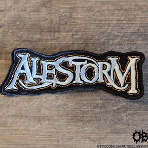 May include: An embroidered fabric patch featuring the word ALESTORM in stylized, light-colored lettering with a dark outline. The text is stitched in a pale, off-white thread against a black background, with a thin orange border surrounding the letters.