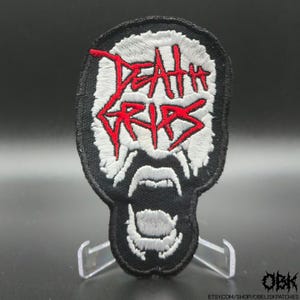 May include: Embroidered patch with the words "Death Grips" in red, stylized lettering. The patch has a black border and a white background, with a design resembling an open mouth. The patch is displayed on a clear stand.