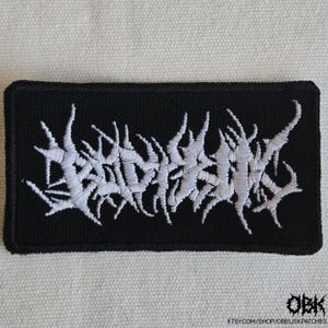 May include: A rectangular black embroidered patch featuring white thread stitched in a stylized, jagged, and illegible death metal band logo font. The design is centered on a solid black fabric background with a thick, stitched black border.