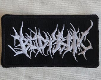 Puede incluir: Un parche rectangular bordado en negro con hilo blanco que forma un logotipo de banda de death metal estilizado, dentado e ilegible. El diseño está centrado sobre un fondo de tela negra sólida con un borde negro grueso y cosido.