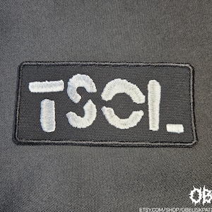 May include: Black rectangular patch with white embroidered text "TSCL." The patch is bordered with black stitching. The background is a dark, textured fabric. The bottom right corner has a white logo and the Etsy shop URL.