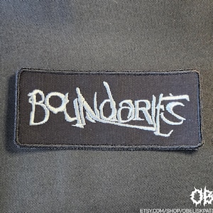 May include: A rectangular black embroidered patch featuring the word Boundaries in a stylized, jagged white font. The patch has a black stitched border and is set against a dark, textured fabric background. The text is clearly legible in white.