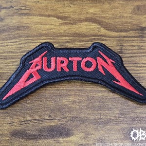 Puede incluir: Parche bordado negro con la palabra "BURTON" en rojo, estilizado con detalles de rayos. El parche tiene una forma única y probablemente está diseñado para ser cosido en ropa o accesorios. El parche está sobre una superficie de madera.